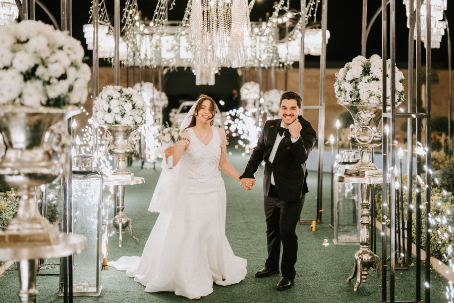 Bride and groom walking hand in hand through an elegant wedding venue with chandeliers, string lights, and white floral arrangements
